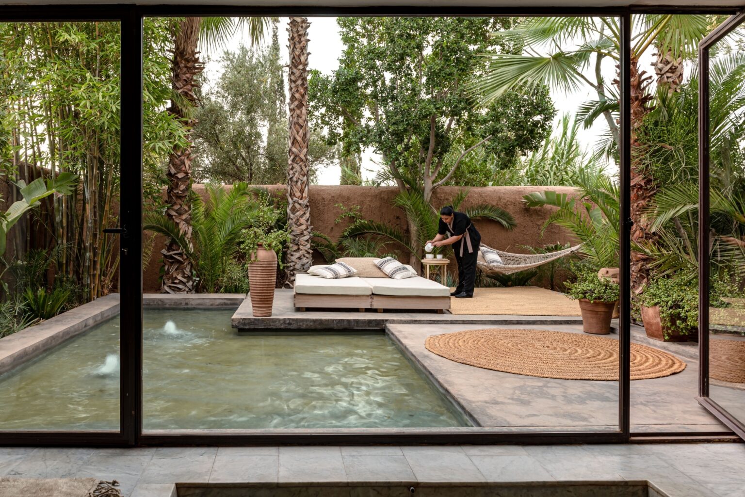 Pool and planted garden of a Marrakech property seen through a steel-framed glass wall, with pisé boundary walls, mature palms and olive trees