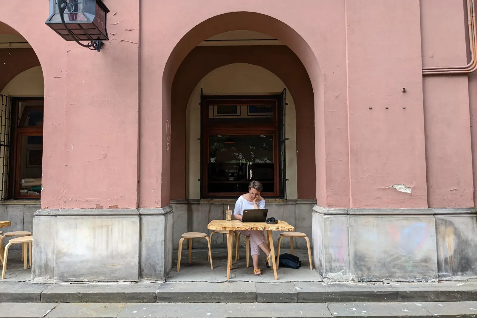 Woman working on a laptop at a café table in front of a pink arched doorway
