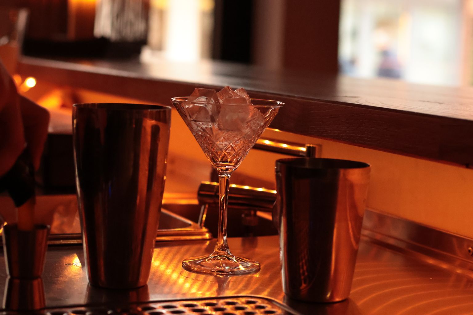 Cut-crystal cocktail glass and steel shakers on a warmly lit bar counter