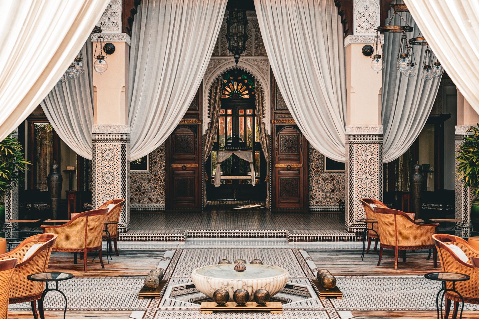 Shaded Moroccan courtyard with zellige tilework, carved cedar doors, white drapes and a marble fountain at its centre