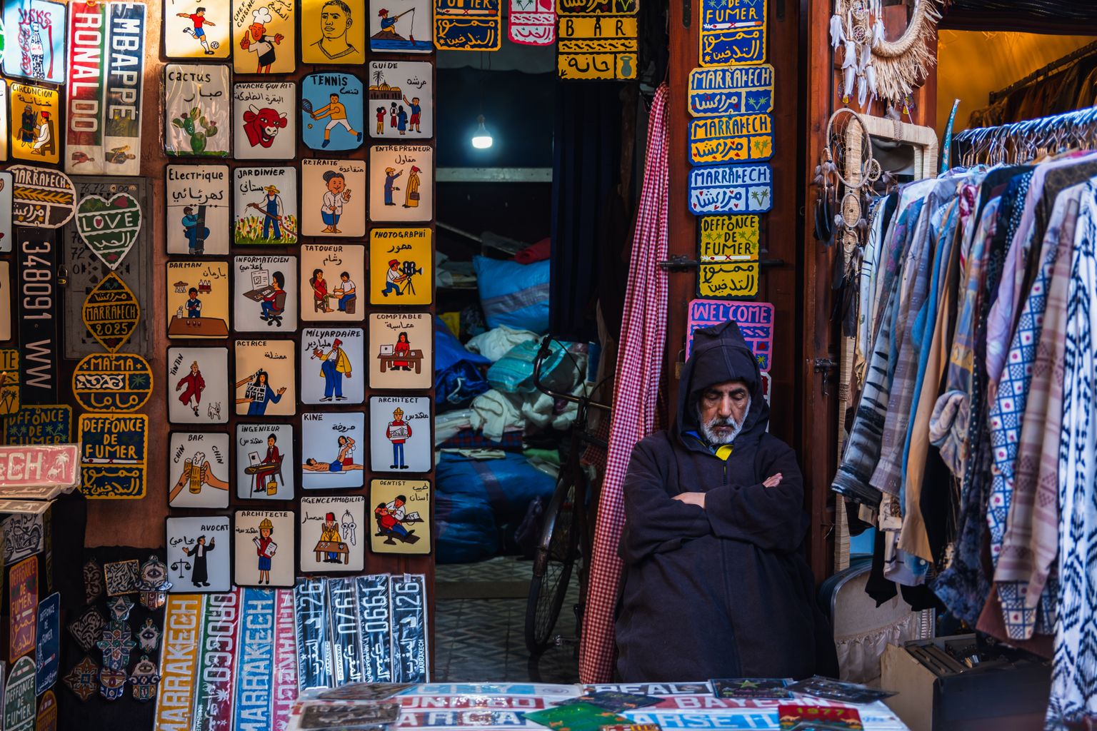 Étal de souk à Marrakech couvert de plaques émaillées peintes à la main portant le nom de la ville en latin et en arabe, à côté d'un portant de vêtements